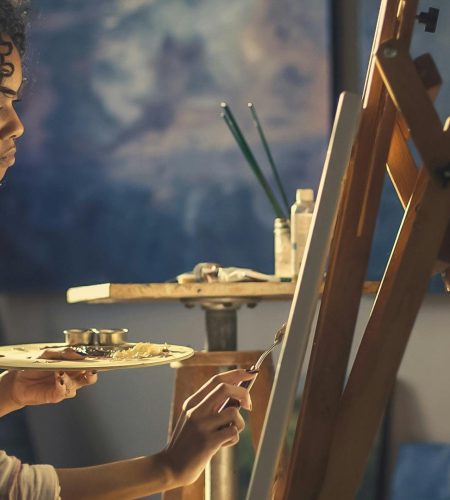 Giới Thiệu Side view of a young woman artist painting on canvas in a cozy studio setting.
